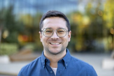 Açık havada gözlük takan gülümseyen bir adamın portresi. Mavi düğmeli bir gömlek giymişti. Sıcak, davetkar bir ifade..