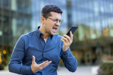 Gözlüklü kızgın bir işadamı dışarıda dikilirken akıllı telefondan bağırıyor. Görünürde hayal kırıklığına uğramış, elleriyle el kol hareketi yapıyor..