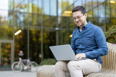 Bir adam bilgisayarını açık havada çalışırken gülümsüyor, modern bir bina ve arka planda bir bisiklet..