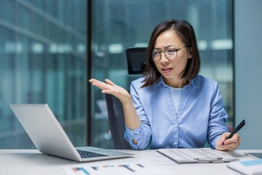 Odaklanmış Asyalı bir iş kadını dizüstü bilgisayarı üzerinde çalışırken şaşkın şaşkın bakar ve bir not defterinin yanında bir kalem tutar..