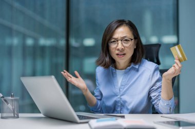 Masasında elinde kredi kartı olan kafası karışmış Asyalı bir kadın. Ofisinde bir dizüstü bilgisayar var..