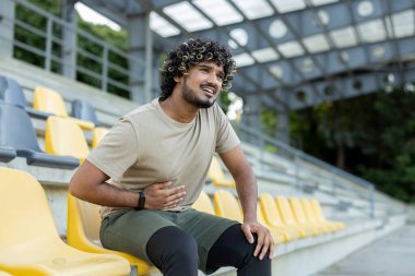 Açık hava antrenmanından sonra stadyum tribünlerinde otururken karın ağrısı çeken genç bir sporcu..