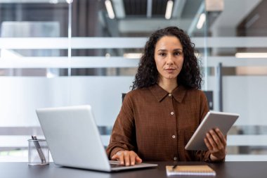 Ofis ortamında iş için laptop ve tablet kullanan ciddi bir iş kadını doğrudan kameraya bakıyor. Odaklanmış ve profesyonel bir resim.