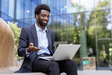 Takım elbiseli neşeli bir Afro-Amerikan işadamı, video görüşmesi sırasında bir bilgisayarla dışarıda, bankta gülümsüyor..