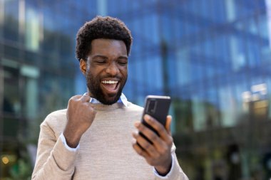 Neşeli bir siyah adam elinde akıllı bir telefonla heyecanla tepki veriyor. Açık havada kazanmayı kutluyor gibi..