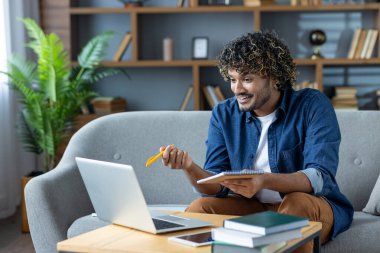 Genç bir adam bir video görüşmesinde dizüstü bilgisayarı ve kalemle koltukta otururken etkileşime geçiyor..