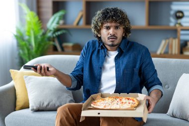 Hayal kırıklığına uğramış bir adam elinde pizza ve telefon ile oturur, muhtemelen teslimattan şikayet etmeye hazırdır. Belli ki mutsuz.