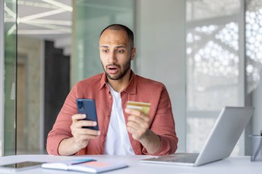 Bir adam telefonuna bakıp kredi kartı tutarken şok oluyor, muhtemelen internetten alışveriş yaparken..