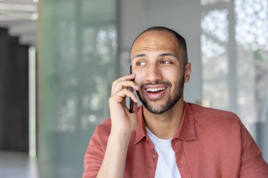 Kırmızı tişörtlü gülümseyen bir adam telefonda, meşgul görünüyor ve mutlu bir ifadeyle başka tarafa bakıyor..