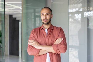 Çapraz kollu kendine güvenen bir adam modern bir ortamda, güçlü ve kendinden emin bir şekilde poz veriyor. Doğal ışık, rahat atmosferi güçlendirir..
