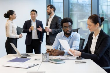 Modern bir ofis ortamında işbirliği yapan çeşitli iş adamları, projeleri için belgeler ve teknolojiye odaklanıyorlar..