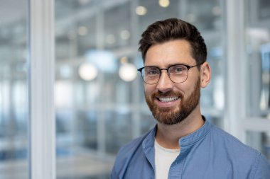 Gözlüklü ve sakallı gülümseyen bir adam modern ofis ortamında bir portre için poz verir..