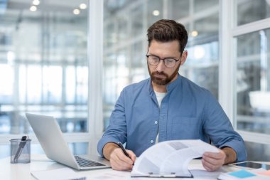 Gözlüklü bir profesyonel. Evrakları inceliyor. Dizüstü bilgisayarı ve parlak bir ofis ortamında kalemle..