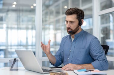 Kulaklık takan sakallı bir adam laptop ekranına odaklanmış, muhtemelen bir iş toplantısında..