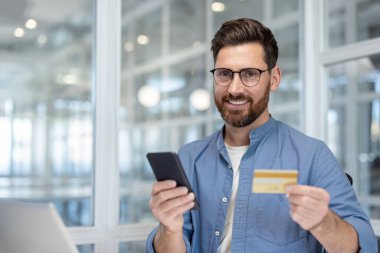 Elinde kredi kartı ve akıllı telefon tutan gülümseyen bir adam, muhtemelen online alışveriş yapıyor parlak bir ofis ortamında.