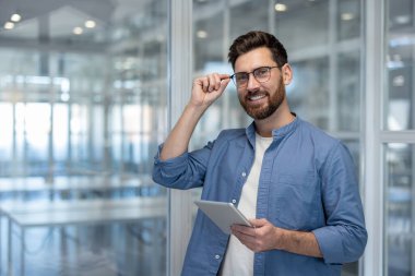 Sakallı, gözlüklü, elinde tablet olan ve gözlüklerine diğer eliyle dokunan gülümseyen bir adam..