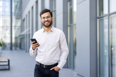 Gülümseyen bir iş adamı, elinde akıllı bir telefon, gözlük ve gömlek, modern bir binanın yanında duruyor..