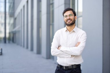 Gözlüklü gülümseyen Hintli bir işadamı dışarıda kollarını kavuşturmuş, güvenini temsil ediyor..