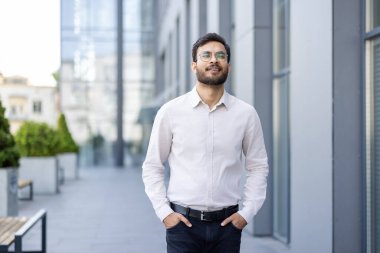 Gözlüklü genç Hintli bir adam, modern bir binanın önünde güvenle duruyor, hafifçe gülümseyerek yukarıya bakıyor..