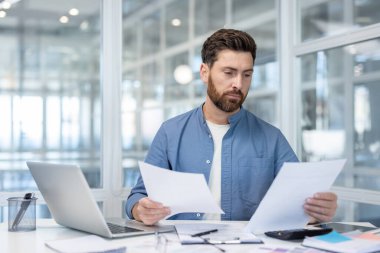 Odaklanmış bir işadamı, ofis gereksinimleriyle çevrili belgelerini masasında gözden geçirir, modern bir çalışma ortamında yakalanır..