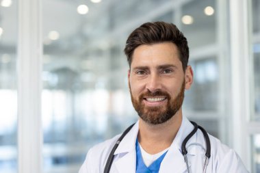 Sakallı yakışıklı bir erkek doktor, steteskop ve laboratuvar önlüğü giyiyor, parlak, temiz bir ofis düzeninde..