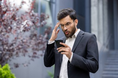 Takım elbiseli bir işadamı telefonuna dışarıda, binanın yakınında bakarken şaşkınlıkla tepki veriyor..