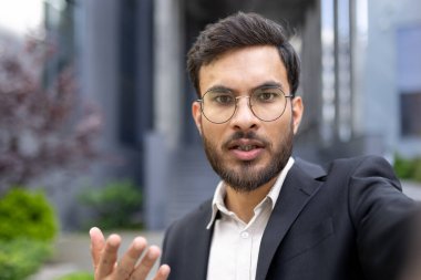 Takım elbiseli bir adam dışarıdaki bir videoda, elleri havada canlı bir şekilde konuşuyor.