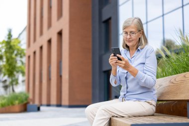 Modern bir binanın yanındaki bir bankta otururken akıllı telefon kullanan gözlüklü yaşlı bir kadın..