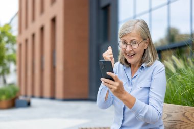 Gözlüklü, mutlu, yaşlı bir kadın telefonunu görüyor ve dışarıda heyecanını dile getiriyor. Bina arka planı sahneye modern bir dokunuş katıyor..