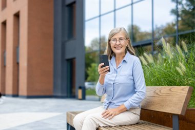 Gülümseyen son sınıf öğrencisi bir kadın bankta oturuyor ve elinde modern bir arkaplan ile akıllı bir telefon tutuyor. Mutlu görünüyor, muhtemelen iletişim kuruyor ya da geziniyor..