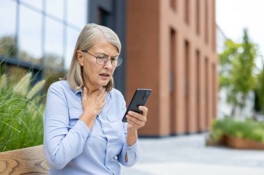 Yaşlı bir kadın, görünürde sıkıntılı, dışarıda telefonunu okurken kötü haberlere tepki veriyor. Şok işareti olarak boğazına dokunuyor.