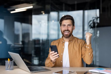 Ofis ortamında akıllı telefon tutarak başarıyı kutlayan mutlu bir işadamı..
