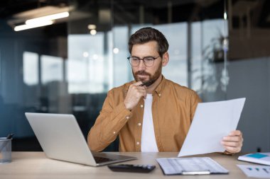 Gözlüklü düşünceli bir işadamı yakınlarda bir laptop ve hesap makinesiyle masasındaki belgeleri inceliyor. Ofis ayarları bulanık bir arkaplan içerir.