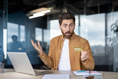 Masada oturan bir adam, bir dizüstü bilgisayarın yanında kredi kartı tutarken şok olmuş gibi görünüyor, muhtemelen mali sorunlar yaşıyor..
