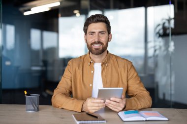 Gülümseyen bir işadamı, modern bir ofis ortamında masasında bir tablet tutuyor..