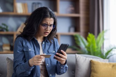 Online hesapla ilgili sorunlar. Evdeki kanepede oturan üzgün Hintli kadın, elinde kredi kartıyla endişeli bir şekilde telefona bakıyor..