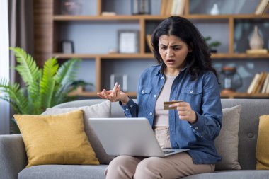 Kafası karışmış ve şok olmuş genç Hintli kadın elinde kredi kartıyla evde oturuyor ve hayal kırıklığına uğramış bir şekilde dizüstü bilgisayar ekranına bakıyor..