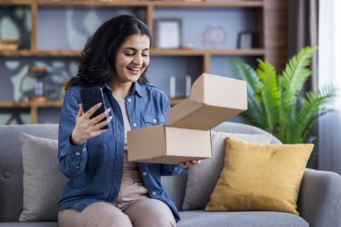 Telefonunu tutarken gülümseyen ve bir teslimat kutusunu açıp heyecanını gösteren bir kadın. Oturma odasındaki rahat bir kanepede oturuyor..