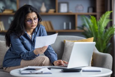 Evdeki kanepede oturan üzgün genç Hintli kadın dizüstü bilgisayar kullanıyor ve hayal kırıklığı içinde evraklara ve belgelere bakıyor..
