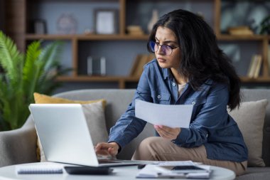 Gözlüklü genç bir kadın evdeki bilgisayarında çalışırken belgelere dikkatlice bakıyor..