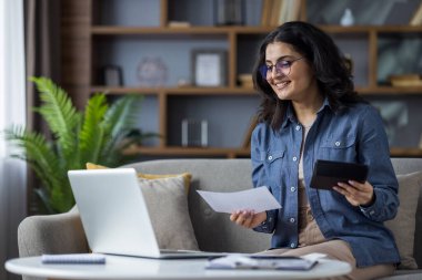 Gülümseyen genç bir kadın evinde çalışır, elinde belgeler ve tabletle kanepede laptopunu kullanır..