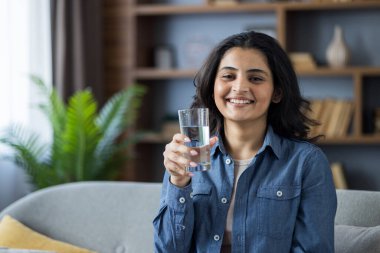 Evinde kanepede oturan, elinde bir bardak temiz su tutan ve kameraya bakan gülümseyen genç bir Hintli kadının yakın plan portresi..
