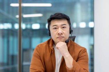 Asyalı bir adam kulaklık takıyor, odaklanmış görünüyor, eli çenesinde, modern bir ofis ortamında..