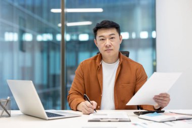 Odaklanmış Asyalı bir adam modern ofis ortamında dokümanları gözden geçirir, doğal ışık ve ciddi bir ifade ile yakalanır..