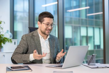 Takım elbiseli, gülümseyen genç bir iş adamı ofisteki iş yerinde oturur ve elleriyle el kol hareketi yaparak bilgisayarla uzaktan konuşur..