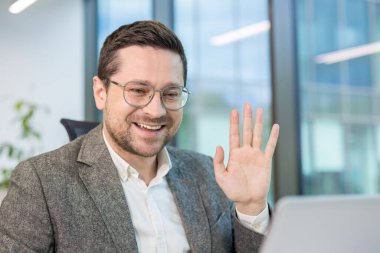 Modern bir ofis ortamında çalışan gülümseyen bir iş adamı, bir video görüşmesi sırasında el sallıyor..