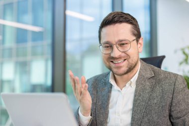 Gülümseyen genç ofis çalışanı çevrimiçi konuşma ve konferans yapıyor. Dizüstü bilgisayarın önünde oturmuş, el kol hareketi yapıyorsun..