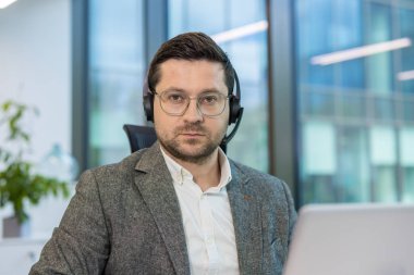 Gri takım elbiseli ve kulaklıklı bir adam doğrudan kameraya bakıyor, muhtemelen müşteri hizmetlerinde ya da çağrı merkezinde çalışıyor..