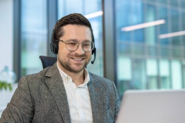 Takım elbiseli ve kulaklıklı genç bir adamın yakın plan fotoğrafı. Ofiste oturuyor ve gülümseyen bir dizüstü bilgisayarla çalışıyor..