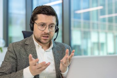 Kulaklık takan bir adam laptopun önünde konuşur, ofiste sanal bir toplantıda görünür..